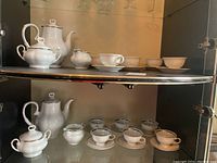 Two porcelain tea sets displayed on glass shelves, showing teapots, sugar bowls, creamers, cups, and saucers.