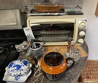 Overall view of the toaster oven with surrounding items including ceramic teapot, sugar bowl, tricker dish, cat-shaped utensil holder, oil bottle, salt and pepper shakers.