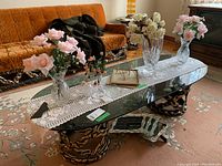 Wide view showing the two crystal vases, artificial flowers in glass vase, picture frames, and glass ashtray on glass coffee table with crocheted runner.