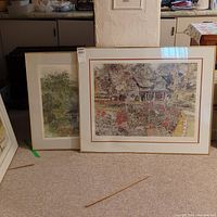 Two framed prints placed on the floor leaning against a wall showing color artwork of a house with garden flowers and a second artwork with greenery.