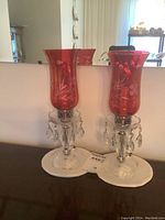 Pair of cranberry etched glass lamps with crystal prisms and clear glass bases shown side by side on surface with mirror background.