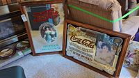 Photo showing both vintage Pepsi Cola and Coca Cola mirrored advertisement signs leaning against a sofa in a room with carpeted flooring. Signs framed in wood with visible wear and age.