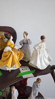 Three Royal Doulton figurines grouped on a dark wood table with mirror background. Visible detail of their elegant dresses and poses.