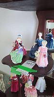 Four Royal Doulton figurines displayed on a multi-tier wooden stand. Various pastel dress colors are visible, and figures show intricate hand-painted detail.