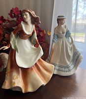 Two Royal Doulton Lady figurines, Autumntime (in the foreground) and Summertime (in the background), shown on a wooden surface with floral decoration behind