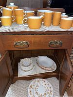 Yellow mugs and cups stacked on wooden table with cream, sugar, and additional cups with white and orange floral plates and bowls below.