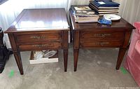 Pair of wooden side tables placed side by side on carpeted floor with various items on top and below. Both tables show warm wood finish, two drawer fronts with ornate brass hardware, and tapered legs with grooves. Light scratches visible on surfaces.
