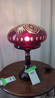 Front and side view of illuminated Bohemian ruby red cut to clear crystal table lamp standing on a round wooden table.