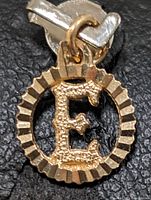 Close-up front view of the gold pendant showing the detailed textured letter 'E' inside a ridged circular frame on a black background.