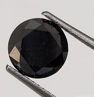 Close-up image of the round cut black diamond gemstone held with tweezers to show size and detail.