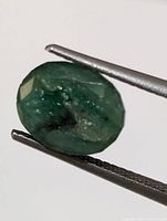 Close-up photo of the oval cut genuine emerald gemstone held by tweezers showing green color and natural inclusions.