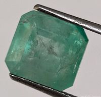 Close-up of genuine emerald green gemstone held by tweezers showing the step cut and translucency.