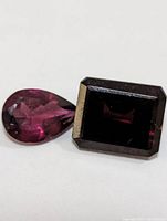 Two genuine gemstones on white background, one teardrop-shaped and one rectangular-cut, both deep reddish-purple color.