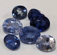 Close-up of seven faceted oval cut sapphires showing color variation from light to dark blue and different sizes.