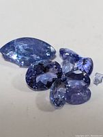 Seven loose tanzanite gemstones in various cuts, colors ranging from blue to violet, one stone broken, all placed on a white background.