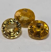 Three genuine yellow sapphire gemstones arranged close together on a white background, showing different hues and round faceted cuts.