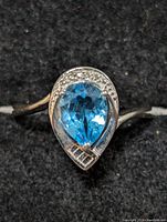 Close-up front view of sterling silver ring with pear-shaped blue topaz and clear accent stones on black background.