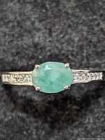 Close-up front view of sterling silver ring featuring oval emerald with four prong setting, clear accent stones on band.