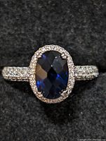 Close-up top view of the sterling silver ring showing the deep blue oval faceted gemstone surrounded by clear stones and a band set with small clear stones.