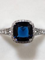 Close-up image showing square cushion-cut blue center stone with halo of small clear stones and silver band with accent stones.