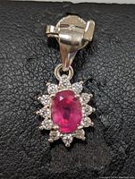 Front view of a sterling silver pendant with oval ruby center stone and clear stone halo on black background.