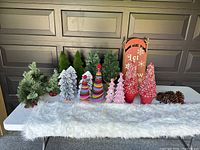 Photo of assorted small Christmas trees including flocked and green evergreens, colorful fabric trees with pom-poms, and pink porcelain trees, along with a red Let it Snow sled, white fluffy table runner, and pinecones all arranged on a white folding table.
