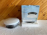 Photo of the Kichler flushmount ceiling light fixture with bronze finish and opaque glass dome next to its original packaging box, clearly showing the box details and the wrapped fixture.