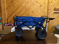 Side view of the folding wagon showing four large heavy duty wheels, blue fabric body, and black metal frame.