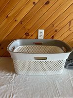White and grey rectangular clothes basket with woven texture and handle slots, placed on a white surface against a wood panel wall background.
