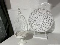 Photo showing a white decorative bird cage and a white carved medallion on stands, both approximately 20 inches high.
