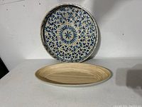 Both items, the round mother-of-pearl and blue tray standing upright against the wall and the long oval light wood and grey bowl below it, placed on a white surface.