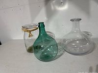 Three glass vases displayed against a white background: clear vase with rope, green bottle-shaped vase, and clear wide-bottom vase.
