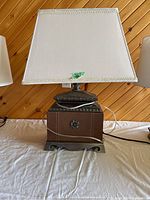 Ornate wooden lamp with drawer, white rectangular fabric shade with fringe, photographed from front on a table against wooden wall background.