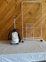 Photo showing Conair clothes steamer on the left and white metal drying rack with six shelves on the right against a wood wall background.