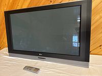 Front view of LG widescreen television with remote control in front on a wooden floor and wood panel wall background.