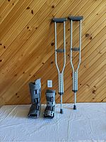 Pair of adjustable aluminum crutches and two grey air casts for left leg/foot placed on white surface in front of a wooden wall background.