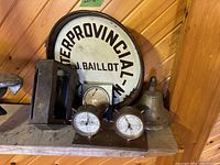 Photo showing a wooden wall shelf holding a round vintage metal sign, a brass nautical bell, a vintage thermometer and barometer mounted on wood, and a vintage fire hose accessory.