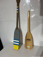 Photo showing two wooden paddles side by side, one shorter with natural wood and branded logo, one longer painted brown with colored stripes and gray/yellow blade