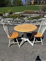 Three pieces of furniture outdoors on asphalt with stone wall and grass background: round pedestal table with removable light wood top and white base, and two matching chairs with white backs and legs and light wood seats.