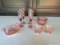 Photo showing 4 pink glass champagne flutes, pink cream and sugar set, decorative bowl, and flower pot on a white surface.