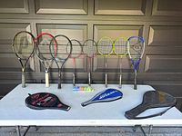 Photo of 3 tennis racquets, 4 badminton racquets with birdies, 1 racquetball or squash racquet, and 3 carrying cases on a table against a garage door background.