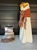 Snowman porch decoration standing upright wearing a yellow hat and orange scarf next to a packaged tree skirt with fur trim.