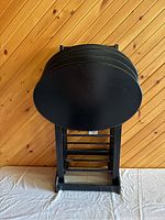 Photo showing 4 ebony black oval folding TV tables stacked on the ebony black stand against wood-paneled wall. Tables have smooth surfaces with minor scratches.