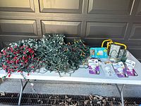 Photo of multiple coiled sets of decorative string lights with red, multicoloured, and white bulbs along with packaged light bulbs and two yellow and black flood lamps on a white table in front of a garage door.