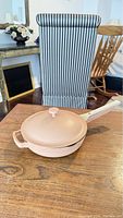 Light pink Always Pan with lid on top placed on wooden table in front of striped chair
