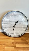 Front view of a large round wall clock with white Roman numerals on a distressed wood background and black decorative hands.