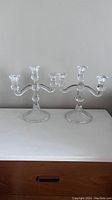 Photo showing two clear crystal candle holders with three arms each on a white surface against a gray background.