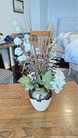 Artificial decorative plant in ceramic vase on wooden table, background shows chair and bedroom