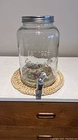 Photo showing a transparent glass drink dispenser with a metal screw-on lid and spout, sitting on top of a large woven coaster.
