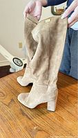 Pair of beige suede knee-high boots being held to show overall profile and shape of the boots with block heels.